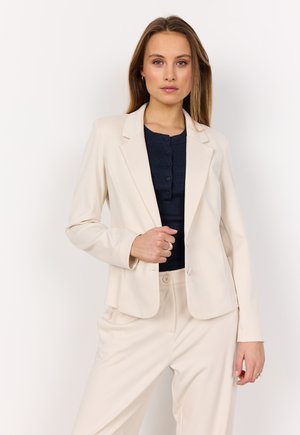 Beige Blazer mit taillierter Passform, Reverskragen und zwei Knöpfen. Getragen über einem dunklen gerippten Oberteil, kombiniert mit passenden beigen Hosen.
