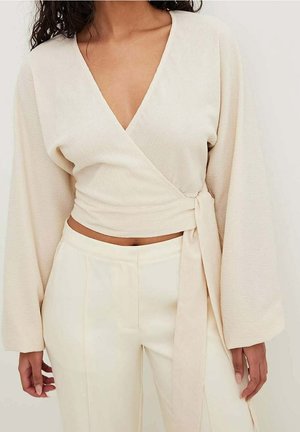 Kobieta ubrana w kremową bluzkę z długimi rękawami z wiązaniem, oraz pasujące spodnie z wysokim stanem, odsłaniające brzuch i ozdobione wiązaniem z boku.