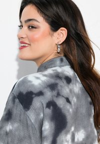 Grijze en witte tie-dye shirt met een kraag en een losse pasvorm, waarbij de textuur zichtbaar is; gedragen met een zilveren oorring en een kleine stud.