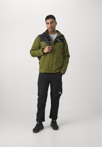 Giacca impermeabile verde con dettagli neri, tasche con zip, pantaloni cargo e scarpe da trekking nere. La giacca ha un collo alto e un cappuccio regolabile.