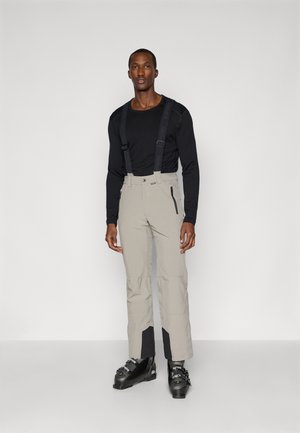 Beige Skihosen mit verstellbaren schwarzen Trägern, zwei Reißverschlusstaschen und schwarzen Akzenten an den Knöcheln. Getragen mit einem langärmeligen schwarzen Shirt und Skischuhen.