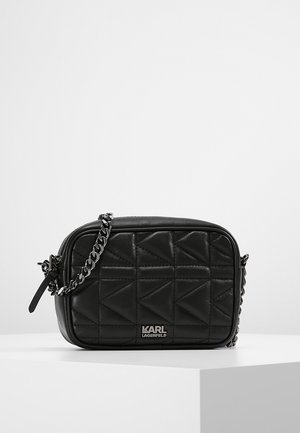 Sac besace en cuir noir matelassé avec bandoulière chaîne argentée et logo KARL LAGERFELD sur le devant, posé sur un piédestal blanc.
