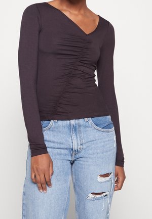 Person iført en mørk langærmet top med rynket V-hals og lyseblå høj taljede revnede jeans mod en ensfarvet baggrund.