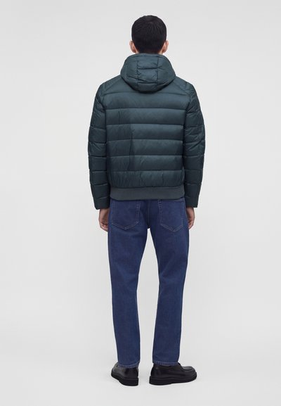 Homme portant une doudoune vert foncé avec capuche, jean bleu et chaussures noires, debout de dos sur un fond clair uni.