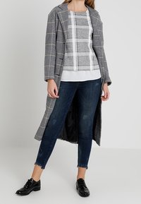 Long manteau à carreaux avec un motif pied-de-poule noir et blanc, assorti à un haut gris à carreaux et un jean skinny foncé. Chaussures noires à lacets.