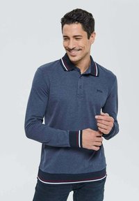 Homme portant un polo à manches longues bleu marine avec un col, des poignets et un ourlet rayés de blanc, rouge et noir, souriant et touchant son poignet.