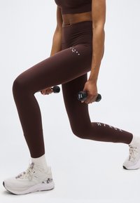 Άτομο που φοράει καφέ αθλητικό κολάν και λευκά αθλητικά παπούτσια, εκτελεί προβολές ενώ κρατάει βαράκια 1 κιλού σε κάθε χέρι.