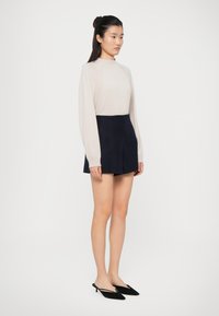 Maglietta lungo beige chiaro, gonna corta blu navy e scarpe con tacco a kitten nere. Tessuti lisci con una silhouette aderente e un colletto alto.