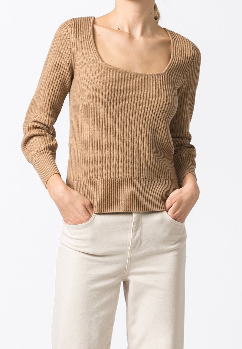 Rippensweater mit quadratischem Ausschnitt in hellbraun, mit enganliegenden langen Ärmeln und weichem Material. Getragen mit hellen Hosen, die Hände in den Taschen.