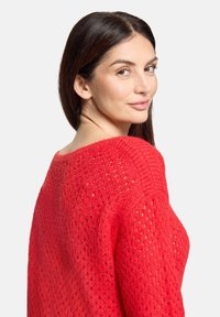 Frau mit langen dunklen Haaren, die einen leuchtend roten, strukturierten Strickpullover trägt und über ihre Schulter blickt, vor einem einfarbigen hellen Hintergrund.