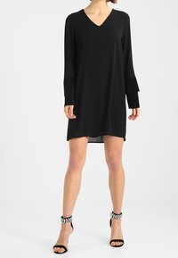 Musta pitkähihaine mekko V-aukolla, virtaavalla siluetilla, rakenteellisilla hihansuilla ja polvimittaisella helmalla, yhdistettynä strappy-mustiin kenkiin, joissa on jalokivikoristelu.