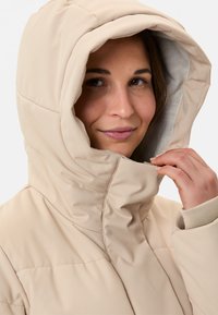 Beige doorgestikte jas met een grote, gestructureerde capuchon, een gladde buitenkant en geribbelde manchetten als accent. Voorzien van een ritssluiting en stikdetails.