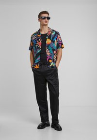 Mand iført sorte solbriller, en farverig blomsteret kortærmet skjorte over en sort tanktop, sorte bukser og sorte sko, står med hænderne i lommerne.