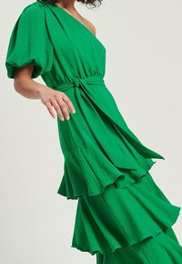 Zelené šaty na jedno rameno vyrobené z texturované látky; mají přiléhavý pas a vrstvenou sukni s volány a uzlem.