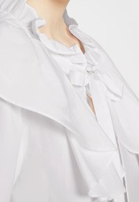 Blouse blanche avec un col et des poignets volantés, ornés de liens en ruban satiné. Le tissu semble lisse et léger, mettant en valeur des textures délicates.