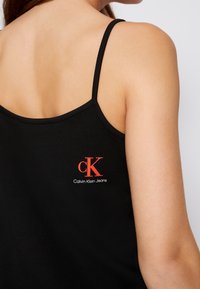 Černý top bez rukávů s tenkými ramínky, na zadní straně s červeným vyšitým logem Calvin Klein Jeans. Hladký, přiléhavý materiál.
