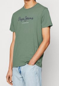Zielona bawełniana koszulka z krótkim rękawem, z ciemnym logo "Pepe Jeans" i napisem "LONDYN" poniżej. Luźny krój i miękka struktura.