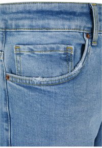 Jeans de mezclilla de color azul claro con un bolsillo desgastado, costura amarilla de contraste y un botón de metal en la cintura. Características de un ajuste estándar.