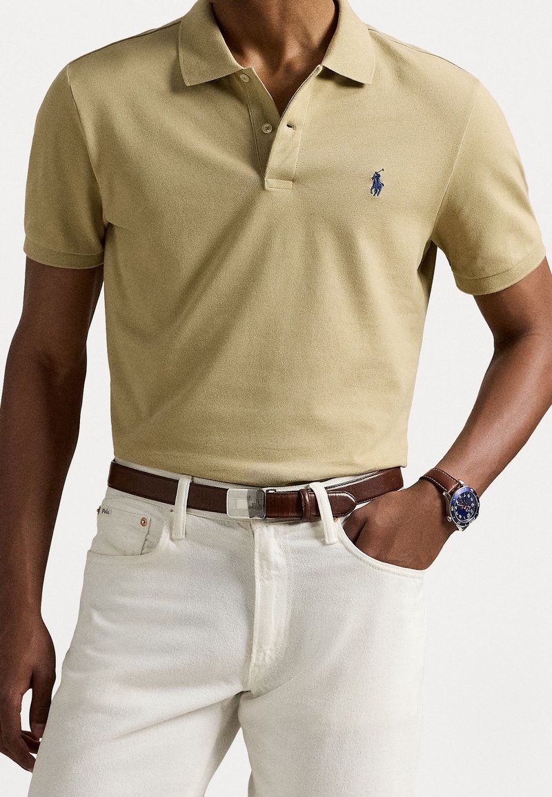 Homme portant un polo beige avec un logo bleu, pantalon blanc, ceinture en cuir marron et montre bleue au poignet, main dans la poche.