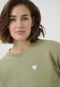 Sweatshirt verde oliva con collo rotondo e piccola grafica di un cuore rosa sulla parte sinistra del petto, realizzato in morbido cotone.
