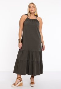 Maxikleid in dunklem olivgrün mit gerafftem Ausschnitt, verstellbaren Trägern und geschichtetem Rüschensaum. Kombiniert mit beigen Keilabsatzsandalen.