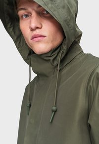 Olivovo zelená nepremokavá bunda s vysokým golierom a nastaviteľnými šnúrkami; má hladkú textúru a zapínanie na cvoky.