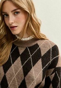 Femme blonde portant un pull en argyle brun et noir avec un col en dentelle à volants, posant devant un fond neutre.