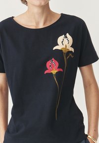 Tamsiai mėlynas medvilninis marškinėliai su laisvu kirpimu, puoštas raudonomis, baltomis ir auksinėmis gėlėmis su kontrastingais juodais akcentais. Trumpomis rankovėmis.
