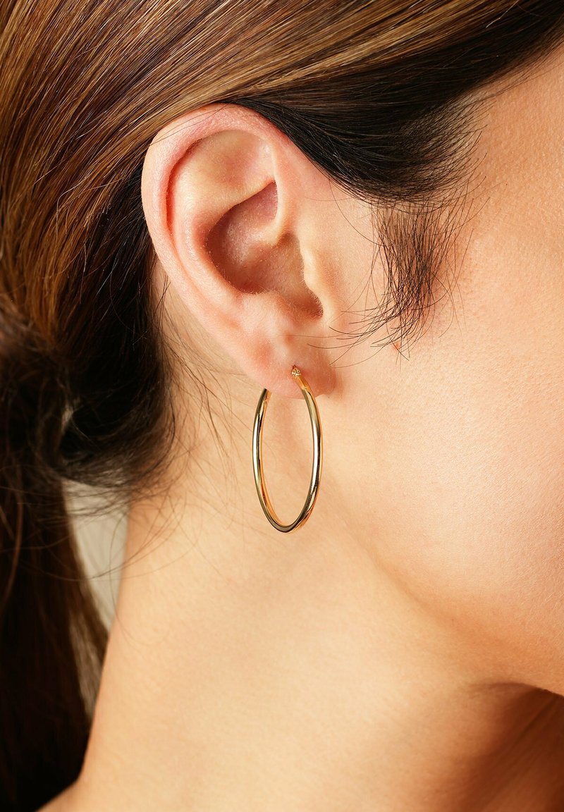 Gros plan sur l'oreille d'une femme avec des cheveux bruns tirés en arrière, portant une boucle d'oreille en or brillant.