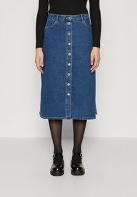 Jeanskjol i blått, knälång med knappknäppning framtill, två sidofickor och en något fransig fåll. Bärs med svarta skor och strumpbyxor.