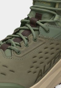 Κοντινό πλάνο πράσινου παπουτσιού πεζοπορίας με επενδυμένη γλώσσα, στρογγυλά κορδόνια, ραμμένες οπές κορδονιών και υφασμένο πέλμα με το εμπορικό σήμα "ALTRA" στο πλάι.