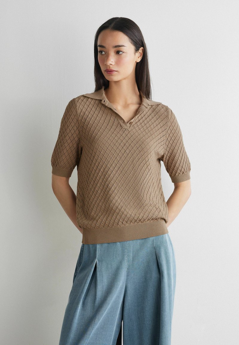 Vrouw met lang donker haar, die een gestructureerde bruine gebreide top met korte mouwen en lichtblauwe broek met wijde pijpen en hoge taille draagt, voor een effen muur.
