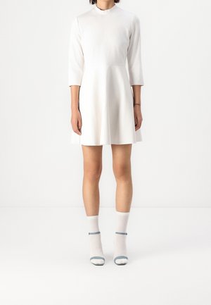 Vrouw draagt een nauwsluitende witte jurk met driekwartmouwen en witte sokken met open-toe sandalen tegen een witte achtergrond.