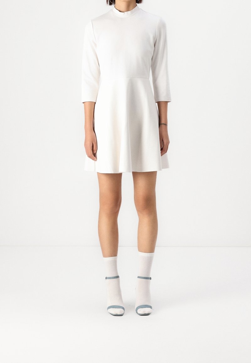Femme portant une robe blanche ajustée avec des manches trois-quarts et des chaussettes blanches avec des sandales ouvertes sur un fond blanc.