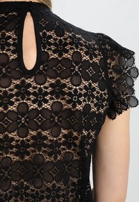 Mustat pitsitoppi, jossa on kukkakuvioita, avonainen kaula-aukko takana ja rypytetyt hihat, jotka korostavat monimutkaista tekstuuria ja muotoilua.