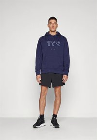 Casaco com capuz azul-marinho com logo "TYR", cordões, bolso frontal; combinado com calções pretos e ténis desportivos pretos. Textura do tecido lisa.