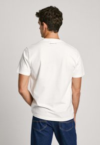 Camiseta de algodón blanca de manga corta y cuello redondo. Presenta una pequeña impresión de "LONDON W11" en la parte superior de la espalda. Combinada con jeans de mezclilla azules.