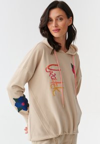 Sudadera beige con cordones, con bordados coloridos y acentos florales en las mangas. Tejido suave, corte holgado y puños acanalados.
