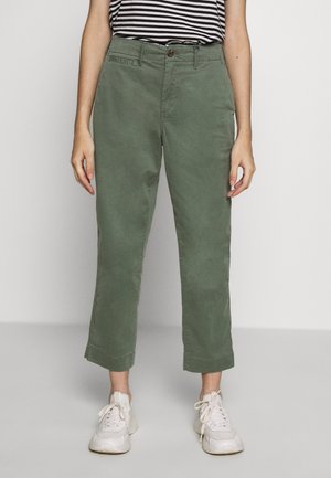 Osoba nosząca podwinięte oliwkowozielone spodnie, białe trampki oraz czarno-białą koszulę w paski, stojąca na tle gładkiej ściany.