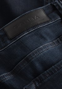 Temno modre kavbojke iz jeansa z gladko teksturo in vidnimi šivi ter črnim usnjenim oznakij z napisom "GABBA" na pasu.