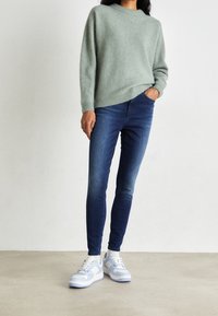 Suéter de punto grueso verde claro con dobladillo acanalado, combinado con jeans ajustados de color azul oscuro. Las zapatillas tienen acentos en azul y blanco.