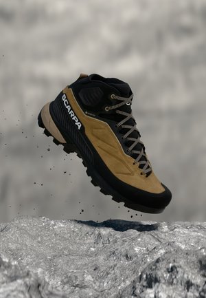 Botte de randonnée Scarpa marron et noire avec Gore-Tex, lacée, en l’air au-dessus d’un terrain rocheux avec des particules de terre qui s’éparpillent.