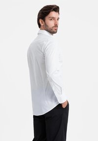 Camisa blanca con pequeños puntos azules, cuello abotonado, mangas largas y un corte entallado, combinada con pantalones negros.