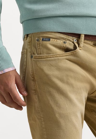 Καφέ βαμβακερές chinos με άνετη εφαρμογή, που διαθέτουν μπροστινή τσέπη, μεταλλικά αξεσουάρ και μια μπλε ετικέτα που γράφει "Polo". Ορατή υφή του υφάσματος.