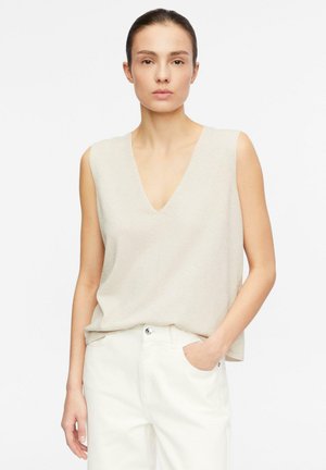 Kobieta o jasnej karnacji z włosami zaczesanymi do tyłu, ubrana w beżowy top bez rękawów z dekoltem w serek oraz białe spodnie, stojąca na jednolitym białym tle.