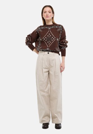 Femme aux cheveux raides debout portant un pull marron à motifs, un pantalon large beige et des chaussures marron foncé sur un fond blanc.