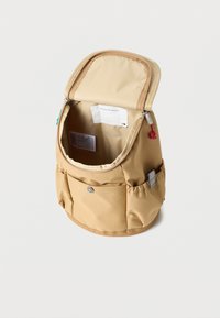 Μικρό μπεζ σακίδιο με ανοιχτό φερμουάρ στην κορυφή, μπροστινή τσέπη με κουμπί, και ελαστικές πλαϊνές τσέπες, πάνω σε απλό φόντο.