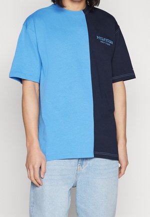 Osoba nosząca krótki rękaw, koszulkę w połowie jasnoniebieską i w połowie granatową z napisem „Hilfiger New York” na piersi, połączoną z jasnoniebieskimi dżinsami.