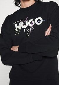 Zwarte sweatshirt met een opvallend wit "HUGO" logo met kleurrijke, artistieke accenten, ronde halslijn en geribbelde manchetten.