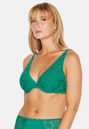 Femme portant un soutien-gorge en dentelle verte et des sous-vêtements assortis, debout devant un fond blanc uni avec une expression neutre.